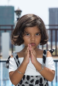 A young child with short dark hair stands with their hands pressed together in a gesture resembling prayer or concentration. The child is wearing a white top with patterned overalls and silver bracelets. Two tall buildings and a lamp post are visible in the blurred background.