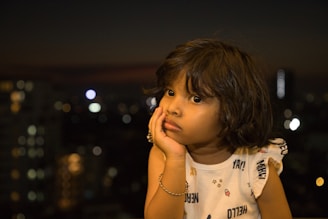 A child with expressive eyes rests their chin on their hand, looking contemplatively towards the right. The background is an out-of-focus cityscape at night, with scattered lights creating a bokeh effect. The child has short dark hair and is wearing a light-colored sleeveless shirt with playful designs.