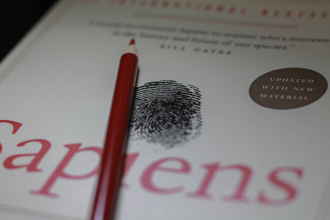 A red pencil rests on the cover of a book titled 'Sapiens'. The cover features a black fingerprint graphic and a round sticker indicating updated material. The text is in red and black.