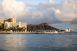 A coastal scene featuring a large waterfront hotel complex with numerous palm trees in the foreground. Behind the buildings, an iconic volcanic crater rises against a backdrop of partly cloudy skies. The calm ocean waters in the foreground reflect the surrounding scenery.