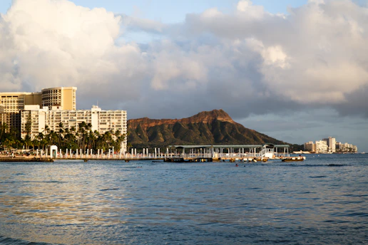 A coastal scene featuring a large waterfront hotel complex with numerous palm trees in the foreground. Behind the buildings, an iconic volcanic crater rises against a backdrop of partly cloudy skies. The calm ocean waters in the foreground reflect the surrounding scenery.