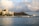 A coastal scene featuring a large waterfront hotel complex with numerous palm trees in the foreground. Behind the buildings, an iconic volcanic crater rises against a backdrop of partly cloudy skies. The calm ocean waters in the foreground reflect the surrounding scenery.