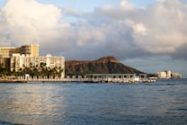 A coastal scene featuring a large waterfront hotel complex with numerous palm trees in the foreground. Behind the buildings, an iconic volcanic crater rises against a backdrop of partly cloudy skies. The calm ocean waters in the foreground reflect the surrounding scenery.