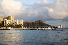 A coastal scene featuring a large waterfront hotel complex with numerous palm trees in the foreground. Behind the buildings, an iconic volcanic crater rises against a backdrop of partly cloudy skies. The calm ocean waters in the foreground reflect the surrounding scenery.