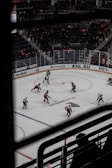 A hockey game taking place in an indoor arena with players wearing red and white uniforms. Spectators can be seen in the background, seated in the stands, and the rink is surrounded by advertisements. The image is taken from an elevated angle through metal railings.