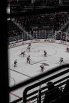 A hockey game taking place in an indoor arena with players wearing red and white uniforms. Spectators can be seen in the background, seated in the stands, and the rink is surrounded by advertisements. The image is taken from an elevated angle through metal railings.
