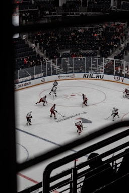 A hockey game taking place in an indoor arena with players wearing red and white uniforms. Spectators can be seen in the background, seated in the stands, and the rink is surrounded by advertisements. The image is taken from an elevated angle through metal railings.