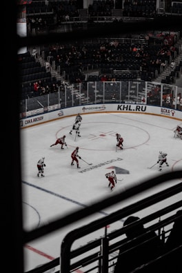 A hockey game taking place in an indoor arena with players wearing red and white uniforms. Spectators can be seen in the background, seated in the stands, and the rink is surrounded by advertisements. The image is taken from an elevated angle through metal railings.