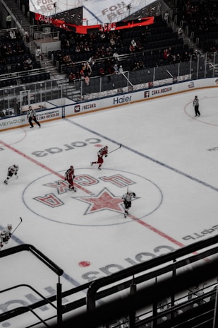 An ice hockey game is underway in an indoor arena with players actively engaged in the sport. Spectators are seated in the stands, while some areas appear sparse. The rink features a large logo at center ice, and electronic advertisements surround the perimeter.