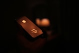 A smartphone is held in a hand against a dark background. The device is illuminated by a warm light source reflecting off its surface, prominently displaying the brand logo. The scene is dimly lit, creating a cozy and intimate atmosphere.