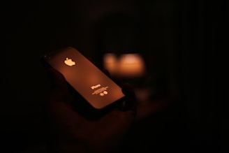 A smartphone is held in a hand against a dark background. The device is illuminated by a warm light source reflecting off its surface, prominently displaying the brand logo. The scene is dimly lit, creating a cozy and intimate atmosphere.