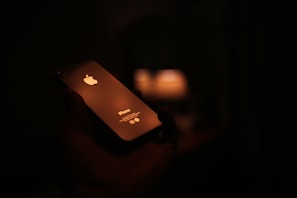 A smartphone is held in a hand against a dark background. The device is illuminated by a warm light source reflecting off its surface, prominently displaying the brand logo. The scene is dimly lit, creating a cozy and intimate atmosphere.