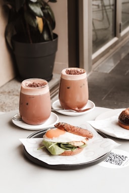 Two glasses of iced chocolate drinks topped with chocolate shavings are placed on white saucers. A sandwich comprising a sunny-side-up egg, fresh spinach leaves, and creamy sauce, held together in a seeded bun, is served on a paper napkin. A potted plant and a glass wall are visible in the background.