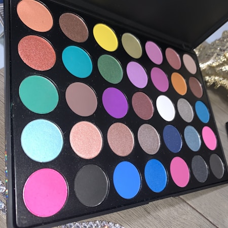 A makeup palette featuring multiple rows of colorful eyeshadow pans in various shades including blue, pink, yellow, green, and neutral tones. The palette is set on a wooden surface with decorative elements in the background.