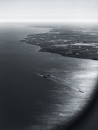 An aerial view of a coastline with a large body of water extending to the horizon. The land features intricate patterns of water channels and patches of greenery, suggesting a marshy or delta region. Several ships are visible in the water, creating light trails on the ocean surface.