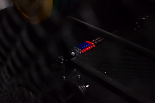 A series of colorful cylindrical objects are aligned neatly on a metal grid. There are red, blue, gray, and copper-colored objects present, with the background partially obscured by a grid or mesh structure.