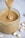 A jar filled with creamy peanut butter, with a spoon lifting a swirl of it from the jar. Whole peanuts are scattered on the gray surface surrounding the jar.