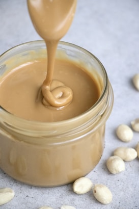 A jar filled with creamy peanut butter, with a spoon lifting a swirl of it from the jar. Whole peanuts are scattered on the gray surface surrounding the jar.