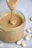 A jar filled with creamy peanut butter, with a spoon lifting a swirl of it from the jar. Whole peanuts are scattered on the gray surface surrounding the jar.