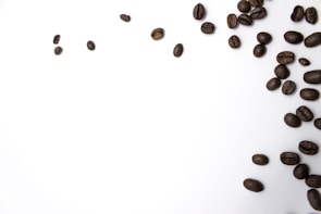 Dark brown coffee beans are scattered on a white surface, predominantly clustered in the bottom right corner, with some beans spread out towards the top left.