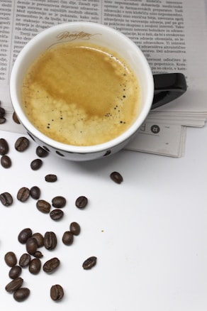 A cup of coffee filled with creamy espresso sits atop an open newspaper. Scattered around the cup are several coffee beans, adding to the overall coffee theme.
