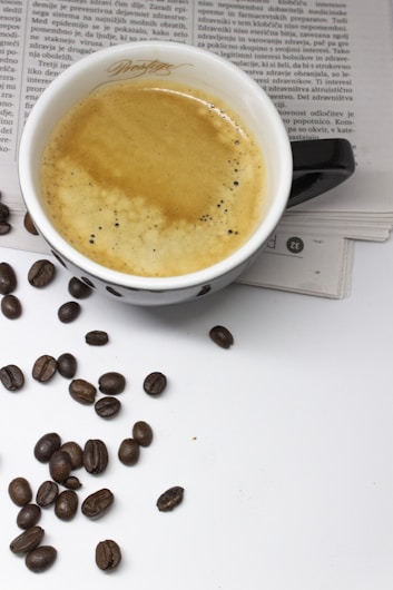 A cup of coffee filled with creamy espresso sits atop an open newspaper. Scattered around the cup are several coffee beans, adding to the overall coffee theme.