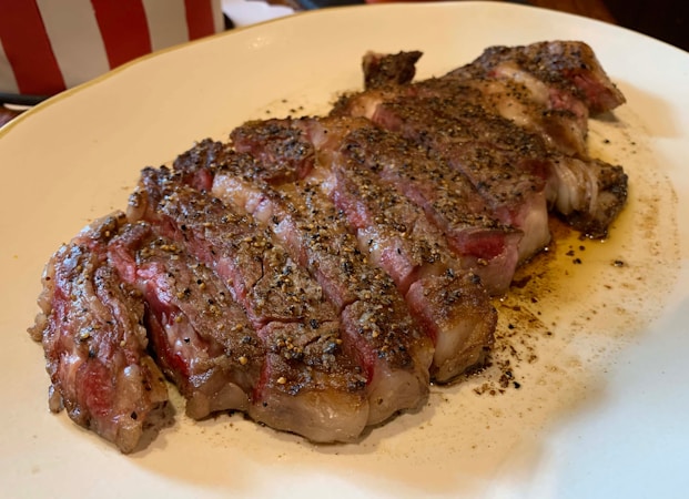 A juicy, medium-rare steak sliced into several pieces is served on a white plate. The steak is well-seasoned with visible black pepper and has a flavorful, seared crust.