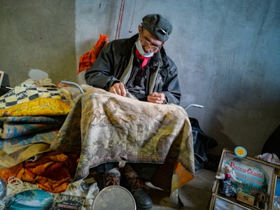 An elderly man, wearing a cap and glasses, sits in a chair working with his hands. He has a face mask hanging around his neck. Various colorful fabrics and handmade items surround him, including a patterned blanket on his lap, and artworks placed on the floor. The setting appears to be indoors, possibly in a workshop or a crafts area, with a grey wall as the backdrop.