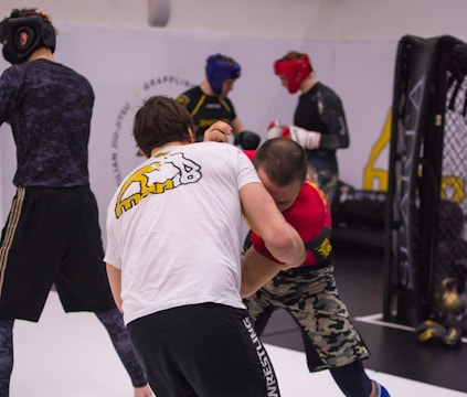 People are engaged in a martial arts training session. Two individuals in the foreground appear to be grappling, wearing protective gear like helmets and gloves. Background participants are also wearing protective gear, practicing various movements. The environment suggests an indoor gym with wrestling mats and a cage-like structure.