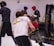 People are engaged in a martial arts training session. Two individuals in the foreground appear to be grappling, wearing protective gear like helmets and gloves. Background participants are also wearing protective gear, practicing various movements. The environment suggests an indoor gym with wrestling mats and a cage-like structure.