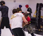 People are engaged in a martial arts training session. Two individuals in the foreground appear to be grappling, wearing protective gear like helmets and gloves. Background participants are also wearing protective gear, practicing various movements. The environment suggests an indoor gym with wrestling mats and a cage-like structure.