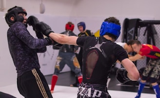 Several individuals are engaged in a sparring session at a gym. They are wearing protective gear including helmets and gloves. One person is landing a punch on another, both wearing headgear and in mid-action poses. The scene is taking place in an indoor combat sports facility, with others also practicing in the background.