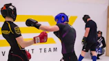 Two individuals are engaged in a sparring session, wearing protective headgear and gloves. One person is delivering a punch while the other has a defensive stance. They are in a training environment with a large logo in the background, and two other individuals are seen in the background, seemingly observing or preparing for their turn.