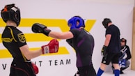 Two individuals are engaged in a sparring session, wearing protective headgear and gloves. One person is delivering a punch while the other has a defensive stance. They are in a training environment with a large logo in the background, and two other individuals are seen in the background, seemingly observing or preparing for their turn.