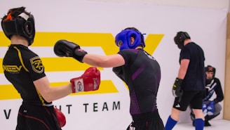 Two individuals are engaged in a sparring session, wearing protective headgear and gloves. One person is delivering a punch while the other has a defensive stance. They are in a training environment with a large logo in the background, and two other individuals are seen in the background, seemingly observing or preparing for their turn.