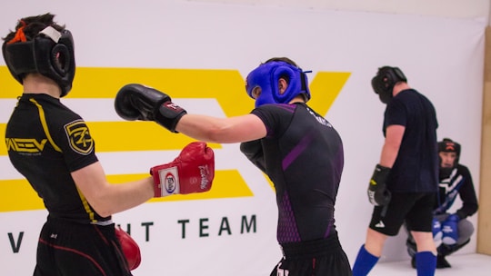 Two individuals are engaged in a sparring session, wearing protective headgear and gloves. One person is delivering a punch while the other has a defensive stance. They are in a training environment with a large logo in the background, and two other individuals are seen in the background, seemingly observing or preparing for their turn.