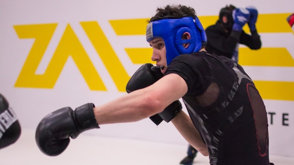 A person wearing a blue headgear and black gloves is engaged in a sparring session, throwing a punch. Another person in protective gear is seen in the background. The setting appears to be a gym or training facility with a white wall and a yellow logo.