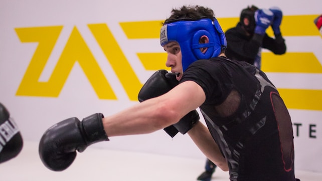 A person wearing a blue headgear and black gloves is engaged in a sparring session, throwing a punch. Another person in protective gear is seen in the background. The setting appears to be a gym or training facility with a white wall and a yellow logo.