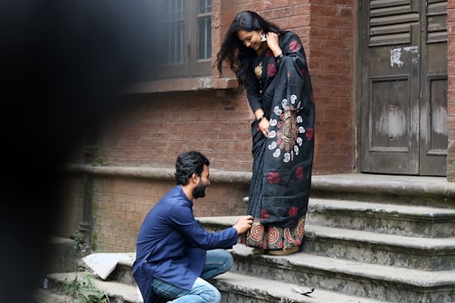 A man kneels on the stone steps of a rustic brick building, adjusting the hem of a woman's beautifully patterned black saree. The woman stands with a joyful expression, her hand touching the side of her face, as she looks down at the man. The scene conveys a sense of care and companionship.