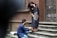 A man kneels on the stone steps of a rustic brick building, adjusting the hem of a woman's beautifully patterned black saree. The woman stands with a joyful expression, her hand touching the side of her face, as she looks down at the man. The scene conveys a sense of care and companionship.
