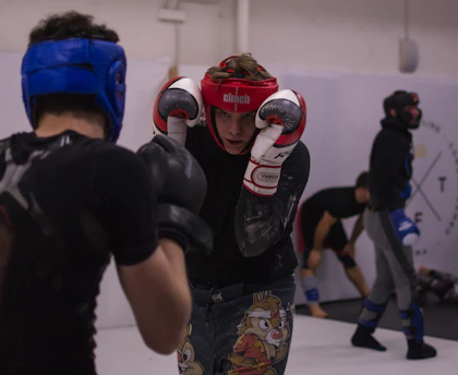 Two individuals engaged in a sparring session, each wearing protective headgear, gloves, and athletic gear. The person in the foreground wears a red helmet and is in a defensive stance, while the person facing them wears a blue helmet. A third person in the background appears to be stretching, possibly preparing to spar.
