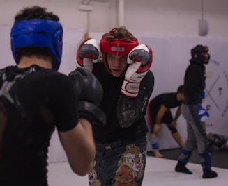 Two individuals engaged in a sparring session, each wearing protective headgear, gloves, and athletic gear. The person in the foreground wears a red helmet and is in a defensive stance, while the person facing them wears a blue helmet. A third person in the background appears to be stretching, possibly preparing to spar.