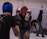 Two individuals engaged in a sparring session, each wearing protective headgear, gloves, and athletic gear. The person in the foreground wears a red helmet and is in a defensive stance, while the person facing them wears a blue helmet. A third person in the background appears to be stretching, possibly preparing to spar.
