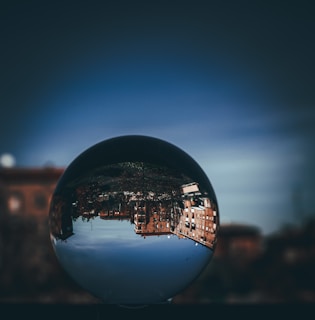 A crystal ball positioned in the foreground reflects an upside-down view of urban buildings and a clear sky. The background is artistically blurred, emphasizing the surreal and artistic nature of the reflection.