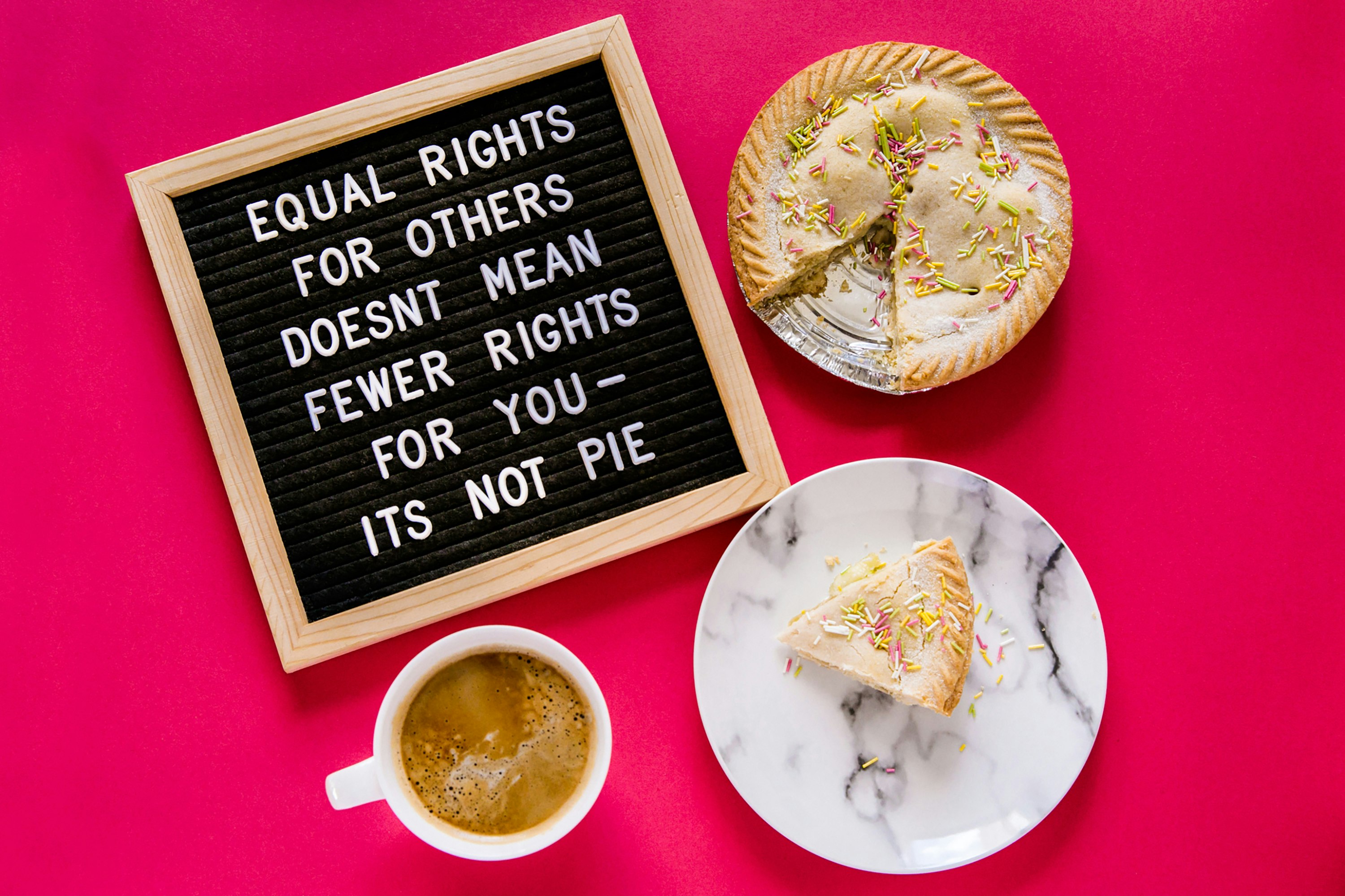 International Women's Day 2021 - Equal rights for others does not mean fewer rights for you, it's not pie. An image from our 2021 Styled Stock collection.

Image is a flat lay from above on a hot pink background. There's a pegboard in the top left showing "Equal rights for others does not mean fewer rights for you - it's not pie" in all caps. The pegboard has a wooden frame and black background with white pegs. In the top right is pie with a slice missing and sprinkles on top. In the bottom right is the slice of pie on a marbled plate. In the bottom left is a white mug filled with coffee.