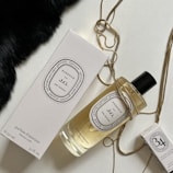 An elegantly arranged display of luxury fragrance items, featuring a large bottle of perfume with a black cap, a matching rectangular box, and a smaller box. The items are adorned with a golden chain draped artfully around them, set against a contrasting background with a patch of black fabric.
