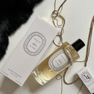 An elegantly arranged display of luxury fragrance items, featuring a large bottle of perfume with a black cap, a matching rectangular box, and a smaller box. The items are adorned with a golden chain draped artfully around them, set against a contrasting background with a patch of black fabric.