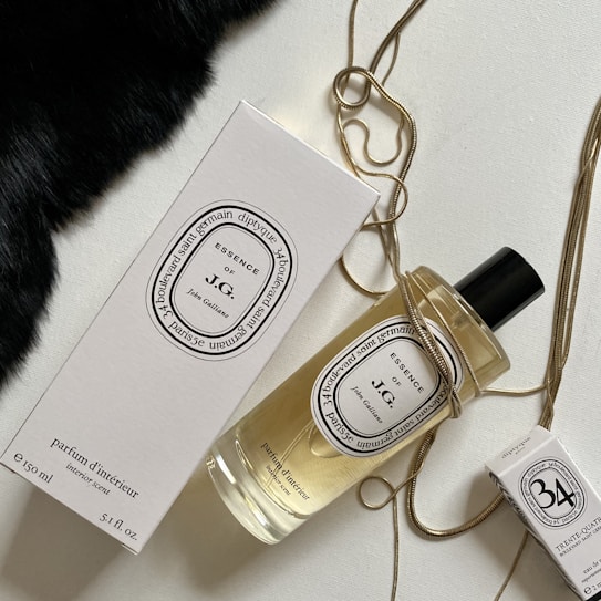 An elegantly arranged display of luxury fragrance items, featuring a large bottle of perfume with a black cap, a matching rectangular box, and a smaller box. The items are adorned with a golden chain draped artfully around them, set against a contrasting background with a patch of black fabric.