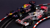 Two model Formula 1 cars are seen racing on a miniature track. The cars are intricately detailed, with one being silver and red, while the other is dark blue with red bull branding. The track features realistic racing elements such as a curve with guardrails and directional markings.