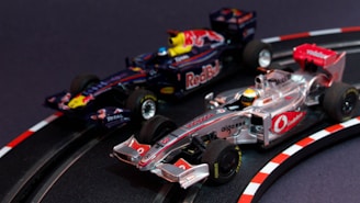 Two model Formula 1 cars are seen racing on a miniature track. The cars are intricately detailed, with one being silver and red, while the other is dark blue with red bull branding. The track features realistic racing elements such as a curve with guardrails and directional markings.